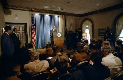 President Nixon at a lecturn against a blue curtain, facing the camera, with the press corps in the foreground facing him.